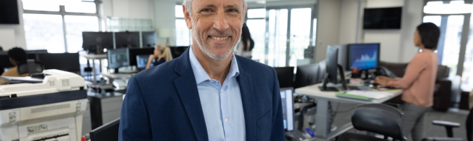   Save Download Preview Portrait of senior Caucasian businessman working in a modern office, standing and smiling to camera, colleagues in the background.