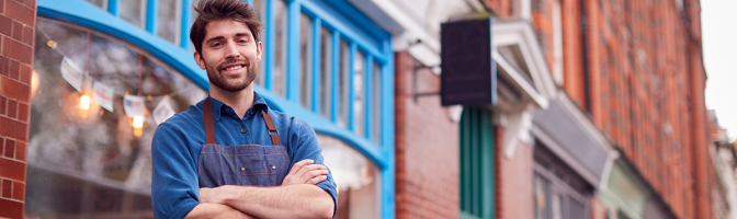 Portrait Of Male Small Business Owner Wearing Apron Standing Outside Shop On Local High Street