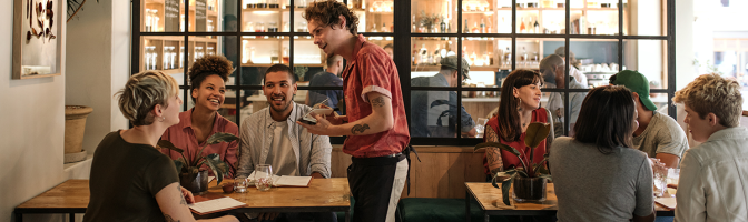 Smiling young waiter taking orders from a diverse group of customers sitting together at a restaurant table