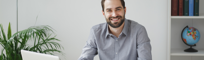 Smiling handsome young bearded business man in gray shirt sitting at desk working on laptop pc computer in light office on white wall background.