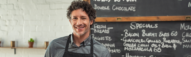 Mature waiter wearing black apron and standing in front of the blackboard with the menu of the day. Portrait of smiling man holding digital tablet and looking at camera.