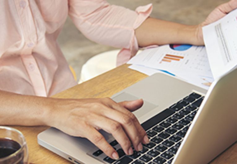 A person checks a print out of data while typing at a laptop.