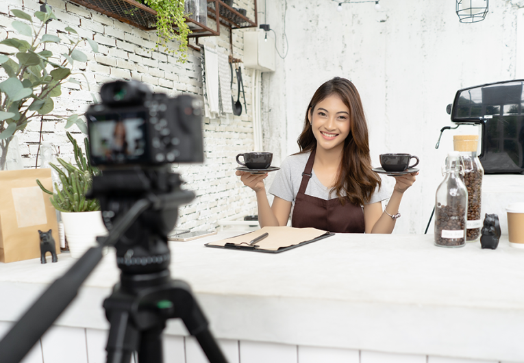 young female barista in apron smiling with recording video camera filming at coffee shop counter.