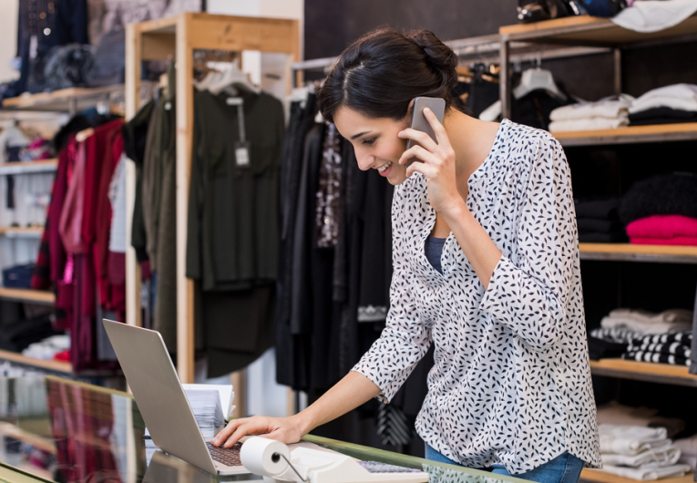Young businesswoman checking laptop and talking on phone while working in her clothing store.