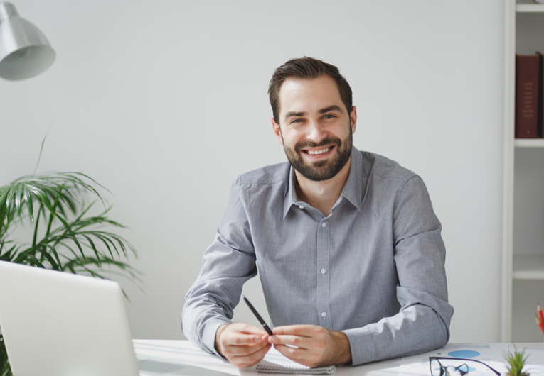 Smiling handsome young bearded business man in gray shirt sitting at desk working on laptop pc computer in light office on white wall background.