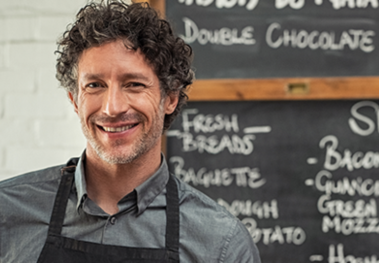 Mature waiter wearing black apron and standing in front of the blackboard with the menu of the day. Portrait of smiling man holding digital tablet and looking at camera.