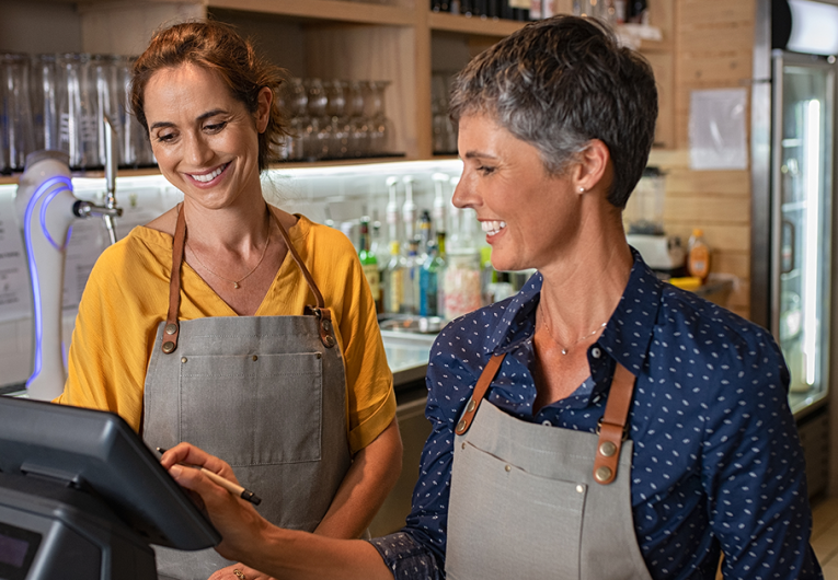 Two happy coffeehouse waitresses in apron smiling and working on cash register.