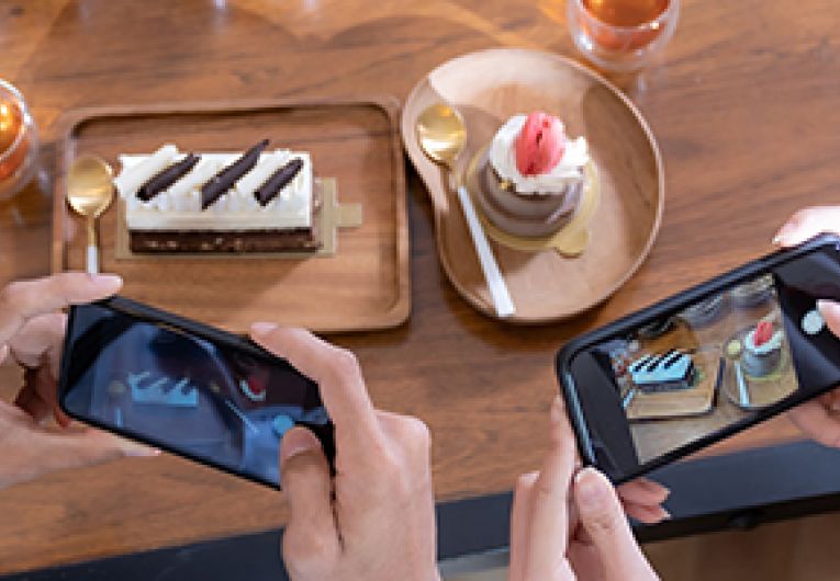 Two people take a photo with their mobile phones of a plate of dessert and beverage. 
