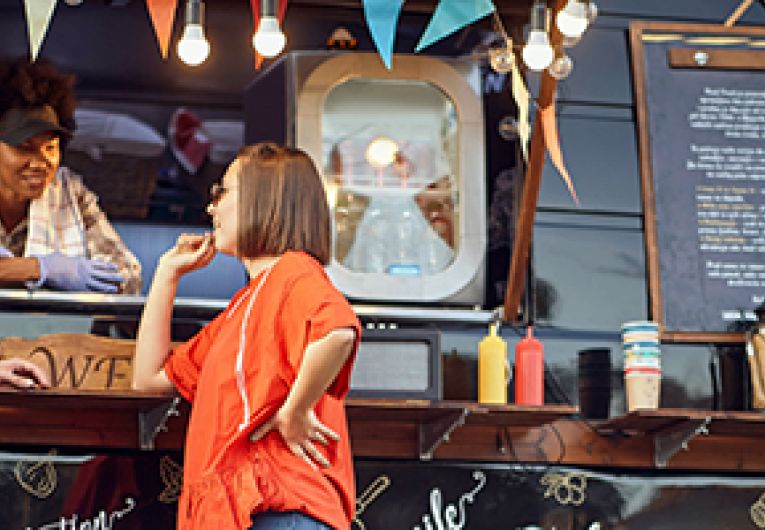 A chef entrepreneur has a friendly conversation with two customers while serving them through the window of her food truck.